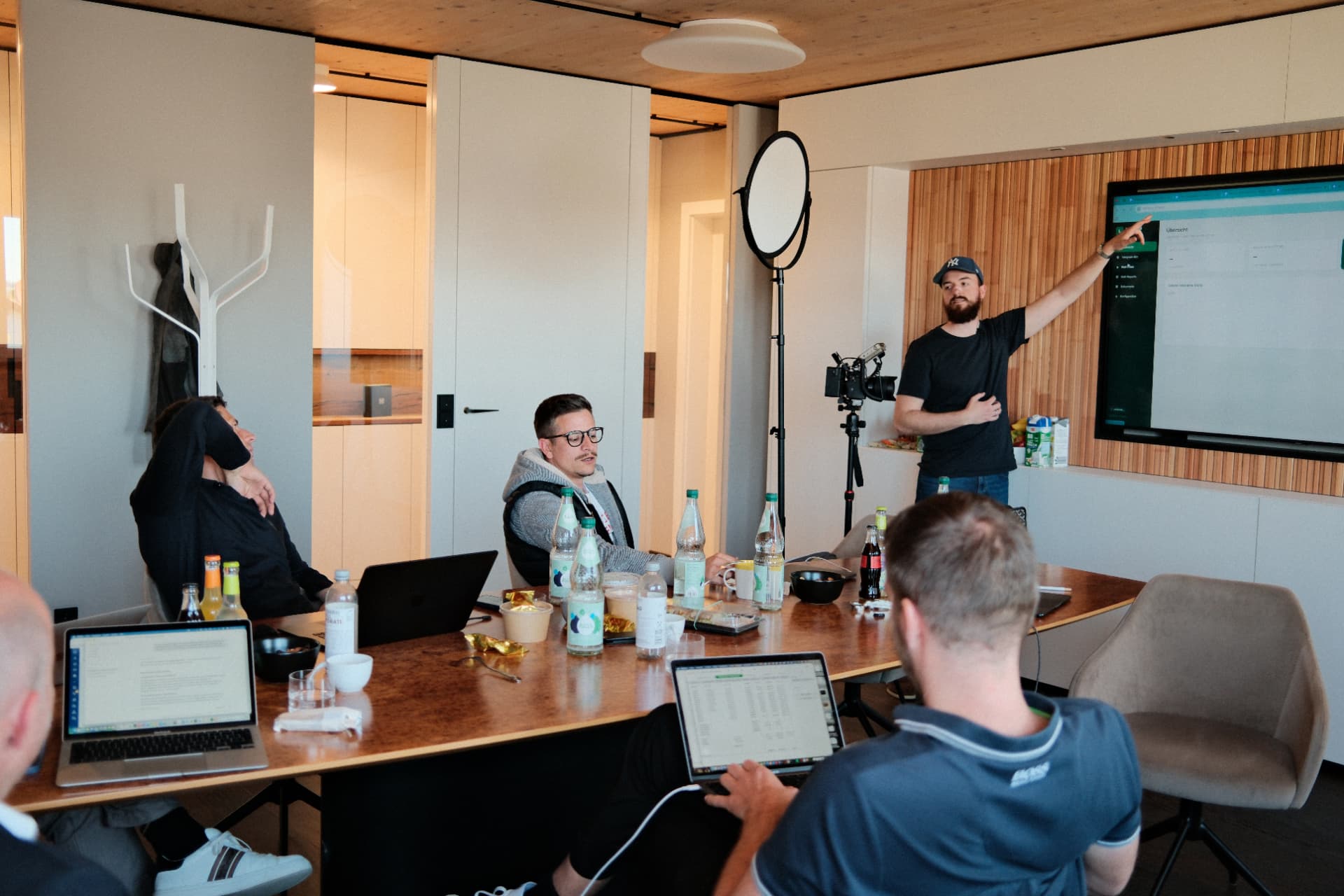 Teilnehmer sitzen am Konferenztisch, Alex präsentiert am Screen